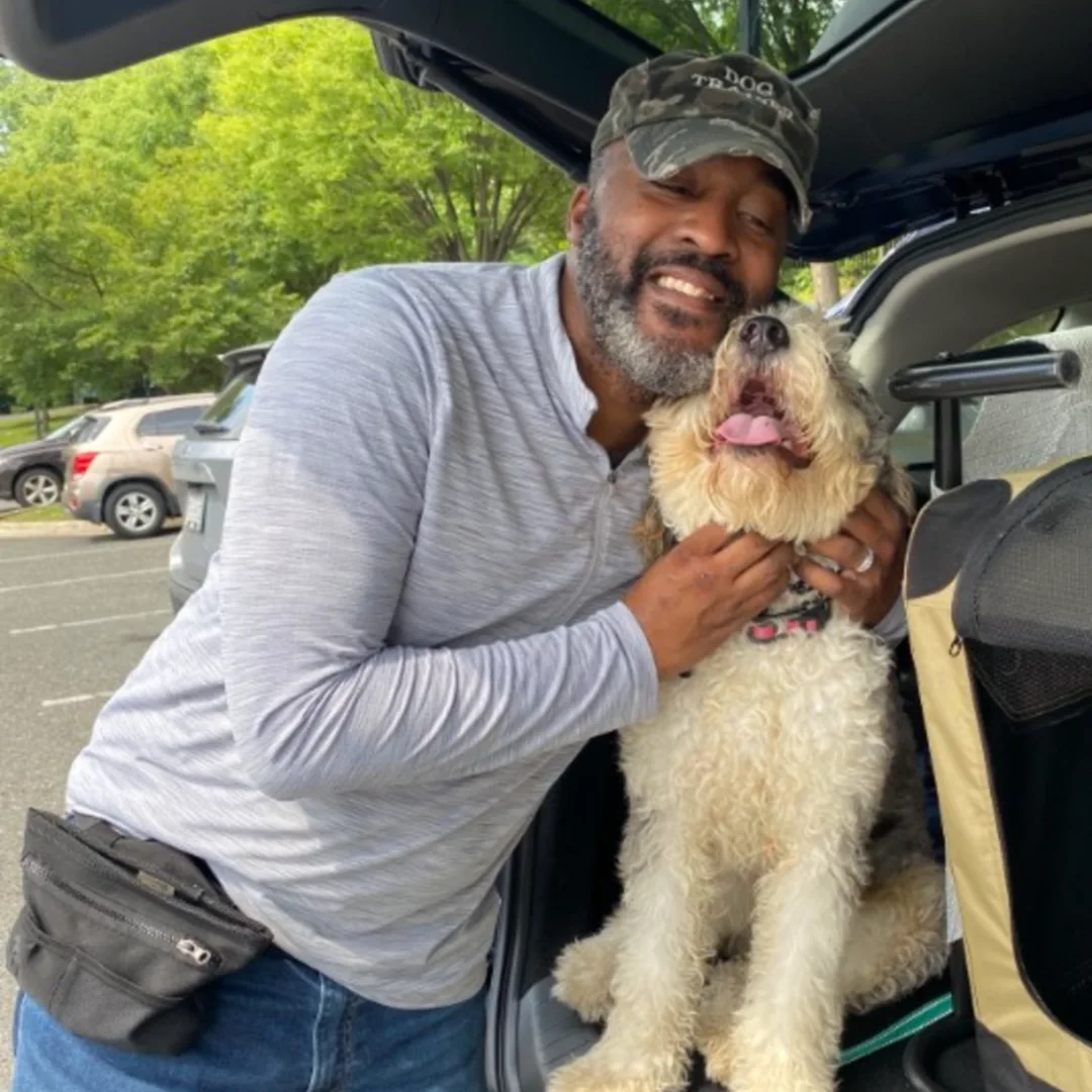 Here's alt text for this image: "A smiling man wearing a camouflage cap and gray long-sleeved shirt kneels on grass next to a white and gray Sheepadoodle with tongue out, in a fenced backyard with stone retaining wall and green shed visible in the background
