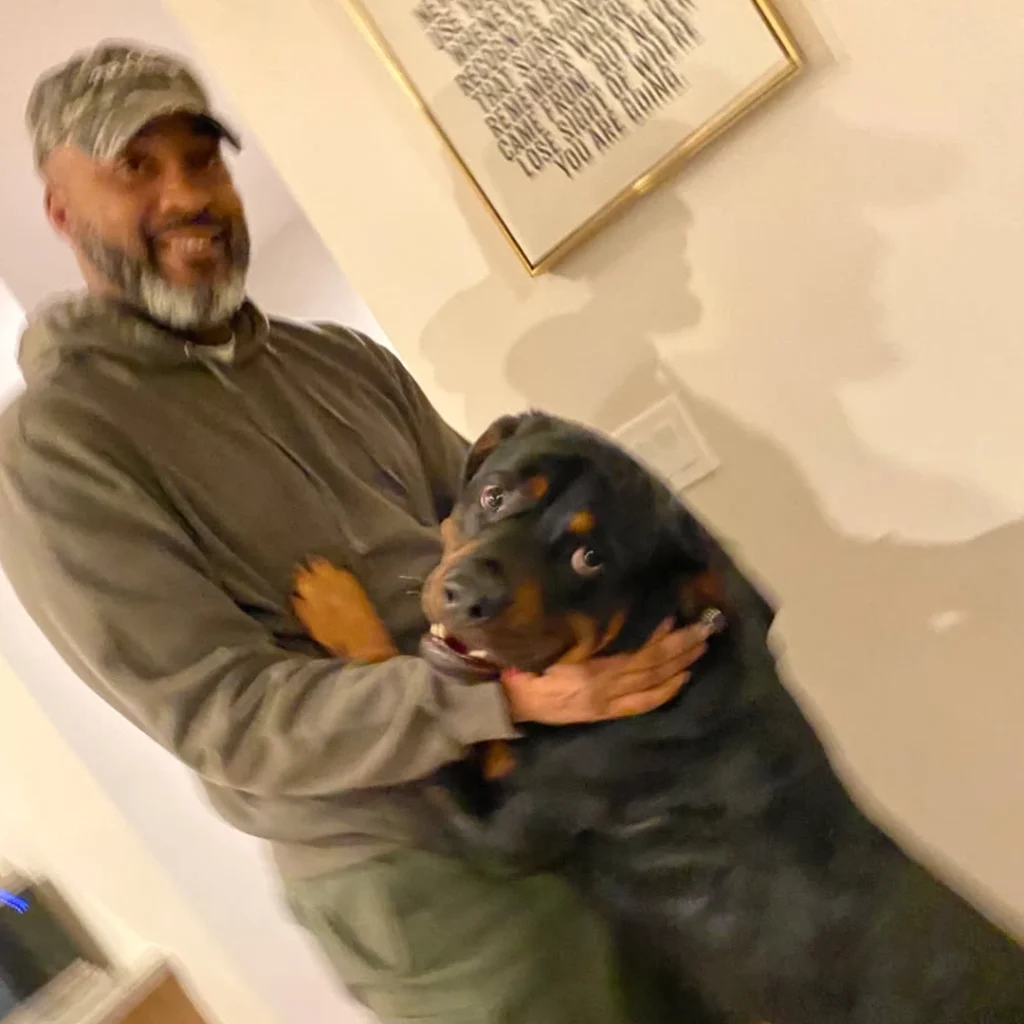 A smiling man wearing a gray cap and olive hoodie holds a black and tan Rottweiler with mouth open in a happy expression, indoors with framed artwork visible on the wall behind them