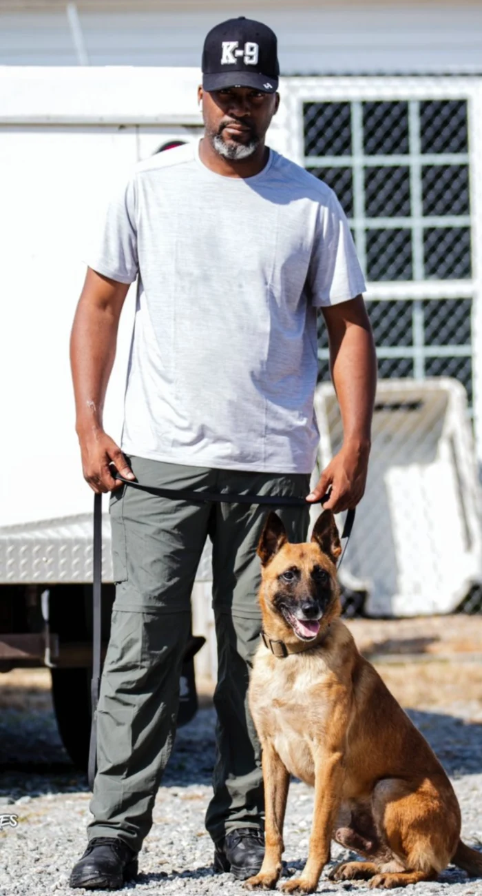 A man wearing a black K-9 cap, white t-shirt, and gray pants stands holding a leash attached to a seated Belgian Malinois with mouth open and tongue out, outdoors in front of a white latticed structure