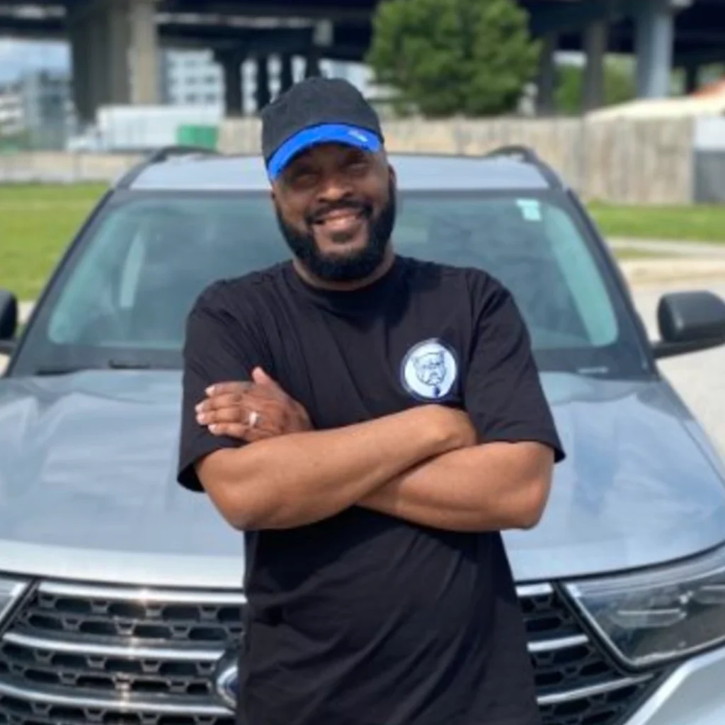 A smiling man with a beard wearing a black cap with blue trim and a black t-shirt with a logo, standing with arms crossed in front of a silver SUV in an outdoor parking area